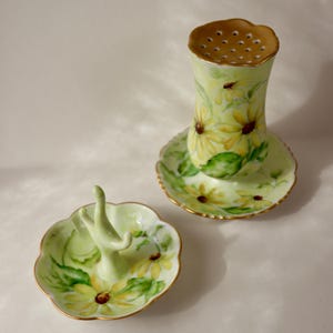 May include: A hand-painted ceramic salt shaker and ring holder set. The salt shaker has a perforated top and sits on a saucer. Both are decorated with yellow daisies and green leaves on a pale green base, with gold edging. A charming floral design.