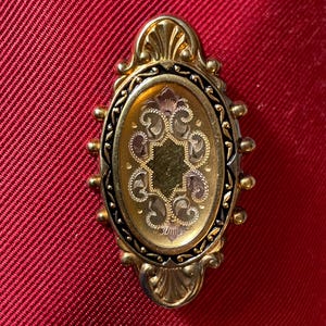 May include: An oval-shaped, gold-toned brooch with an ornate design. The brooch features a central oval with an engraved floral pattern in gold and silver tones, framed by a black border and a decorative gold edge. The background is a textured red fabric.
