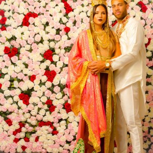 May include: A couple wearing traditional Indian clothing, standing in front of a wall of white and red roses. The woman is wearing a pink and gold sari with a green and gold border. The man is wearing a white shirt and pants with a gold crown.