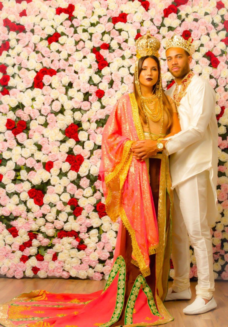 May include: A couple wearing traditional Indian clothing, standing in front of a wall of white and red roses. The woman is wearing a pink and gold sari with a green and gold border. The man is wearing a white shirt and pants with a gold crown.