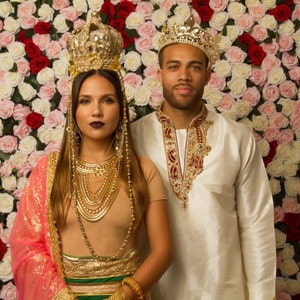 May include: A couple wearing gold crowns and traditional Indian clothing stand in front of a wall of pink and white roses.