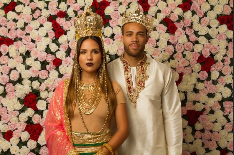 May include: A couple wearing gold crowns and traditional Indian clothing stand in front of a wall of pink and white roses.