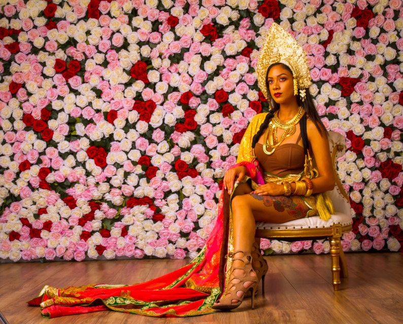 May include: A woman wearing a gold headpiece and a brown top sits on a gold chair. She is wearing gold jewelry and a red and gold patterned shawl. The background is a wall of pink and white roses.