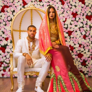 May include: A couple poses for a wedding portrait in front of a wall of pink and white roses. The woman wears a pink and brown sari with gold embroidery and a matching head scarf. The man wears a white suit with gold embroidery. They are sitting on a white throne chair with gold accents.