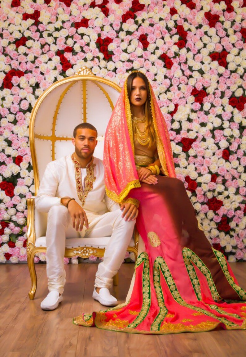 May include: A couple poses for a wedding portrait in front of a wall of pink and white roses. The woman wears a pink and brown sari with gold embroidery and a matching head scarf. The man wears a white suit with gold embroidery. They are sitting on a white throne chair with gold accents.