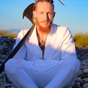 Könnte beinhalten: Ein Mann mit roten Dreadlocks sitzt auf einem Steinhaufen und trägt ein weißes Hemd und eine weiße Hose. Er hält ein großes, dunkles Objekt über seiner Schulter.