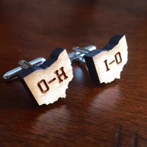 May include: A pair of wooden cufflinks shaped like the state of Ohio. The cufflinks are engraved with "O-H" and "I-O".