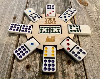 Puede incluir: Un juego de dominó de madera con una placa de nombre personalizada que dice "YOUR NAME". El juego incluye un diseño de locomotora y ocho fichas de dominó con puntos de diferentes colores.