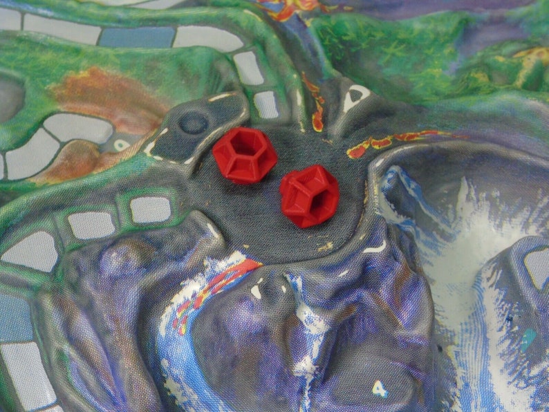 May include: Two red, geometric dice with a hexagonal shape are on a colorful game board.
