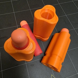 May include: Orange 3D-printed toy parts, including a cylindrical piece, a rounded piece, and a rectangular piece. A red foam dart is also present. The objects are arranged on a black surface with yellow warning symbols.