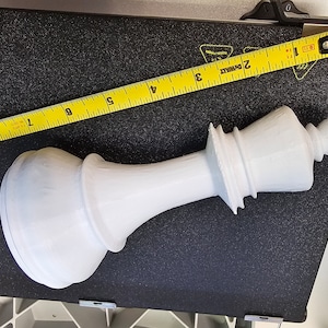 May include: A large white plastic chess piece, a queen, is lying on a black surface. A yellow measuring tape is partially visible in the background, with the numbers 2, 3, 4, 5, 6, 7, 8, and 9 visible.