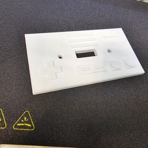 May include: A white plastic wall plate with a Nintendo design. The plate has a power outlet, a light switch, and a series of buttons that resemble the controls on a classic Nintendo Entertainment System controller. The text "Nintendo" is printed on the plate.