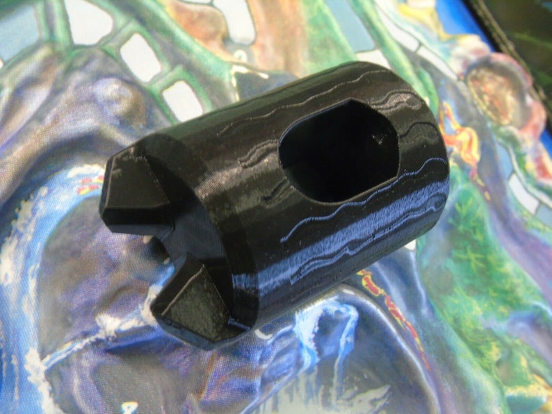 May include: A black plastic 3D printed object with a hexagonal shape and a hole in the center. The object has a textured surface.