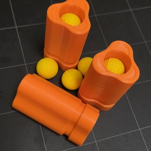 May include: Orange 3D-printed containers with a unique shape, each holding a yellow golf ball. Several additional yellow golf balls are scattered nearby. The objects are arranged on a dark, tiled surface.