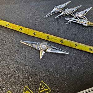 May include: A silver-colored metal star-shaped object with a clear rhinestone in the center. The object is about 5 inches long and is lying on a black surface next to a yellow measuring tape.