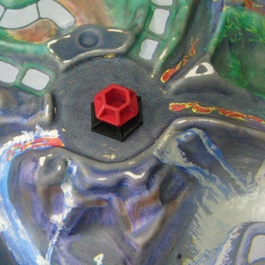 May include: A red and black hexagonal game piece with a raised design. The piece is sitting on a blue and green fabric surface.