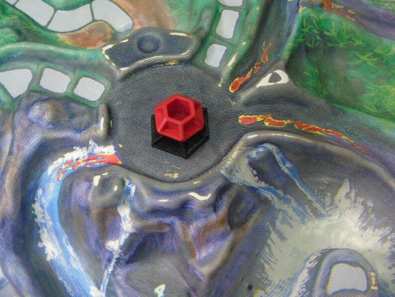 May include: A red and black hexagonal game piece with a raised design. The piece is sitting on a blue and green fabric surface.
