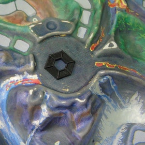 May include: A black hexagonal-shaped plastic piece with a textured surface. The piece is sitting on a colourful fabric background.