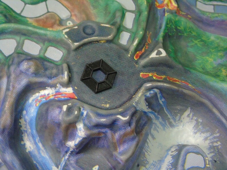 May include: A black hexagonal-shaped plastic piece with a textured surface. The piece is sitting on a colourful fabric background.