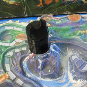 May include: A black plastic game piece shaped like a volcano with a hole in the top. The piece is sitting on a colorful game board with a blue, green, and white design.