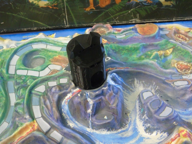 May include: A black plastic game piece shaped like a volcano with a hole in the top. The piece is sitting on a colorful game board with a blue, green, and white design.