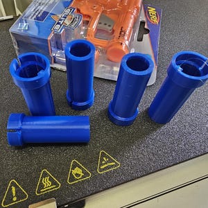 May include: Five blue plastic 3D printed tubes, likely for a Nerf gun, on a black surface. The tubes are different lengths and have a smooth finish. A Nerf Elite Jolt blaster is visible in the background.