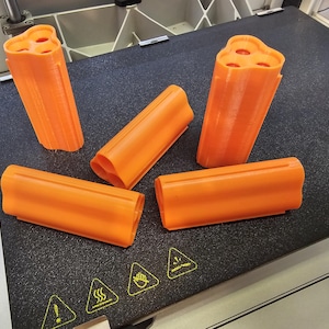 May include: Four orange 3D printed objects, two are cylindrical with three holes in the top, and two are rectangular with rounded edges.