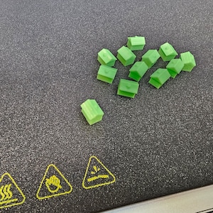 May include: A group of small, green, 3D printed houses on a black surface. The houses are arranged in a cluster, with one house slightly separated from the group.