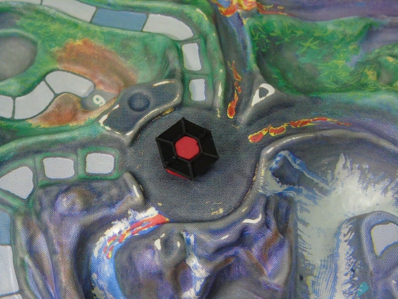May include: A black hexagonal game piece with a red centre, sitting on a blue and purple game board.