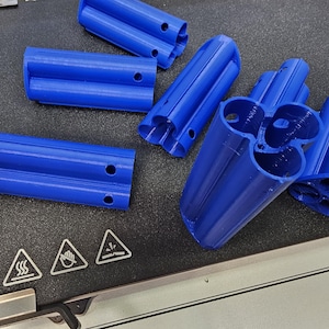 May include: Several bright blue 3D-printed plastic components are arranged on a dark surface. The pieces have a cylindrical shape with multiple holes. Warning symbols are visible in the lower left corner.