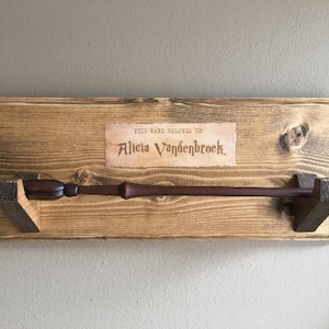 May include: A wooden wand holder with a brown wooden wand resting on it. The holder has a sign that reads "This wand belongs to Alicia Vandenbroek".