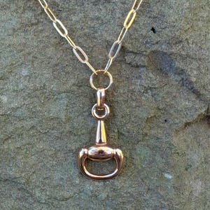 May include: A gold-colored necklace featuring a horse bit pendant. The pendant is a detailed miniature of a horse bit, with a rounded design and a ring at the top. The chain is a delicate link style, and the necklace is displayed against a textured background.