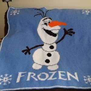Puede incluir: Una manta de crochet azul con un borde blanco que presenta a Olaf, el mu&ntilde;eco de nieve de la pel&iacute;cula Frozen. La manta tiene la palabra "FROZEN" en letras blancas.