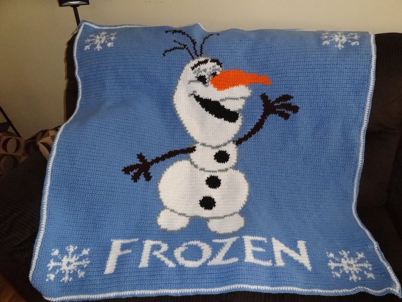 Puede incluir: Una manta de crochet azul con un borde blanco que presenta a Olaf, el mu&ntilde;eco de nieve de la pel&iacute;cula Frozen. La manta tiene la palabra "FROZEN" en letras blancas.