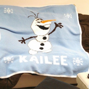Puede incluir: Una manta de crochet azul con un borde blanco presenta una imagen de dibujos animados de Olaf de la pel&iacute;cula Frozen. La manta tiene el nombre "Kailee" bordado en blanco.