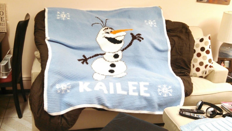 Puede incluir: Una manta de crochet azul con un borde blanco presenta una imagen de dibujos animados de Olaf de la pel&iacute;cula Frozen. La manta tiene el nombre "Kailee" bordado en blanco.