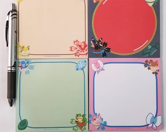 Op de afbeelding: Vier vierkante notitieblokken met kleurrijke randen en hoekillustraties. De blokken hebben verschillende achtergronden, waaronder beige, rood, groen en roze. Een zilveren pen bevindt zich links van de notitieblokken. Elk notitieblok heeft een ander ontwerp.