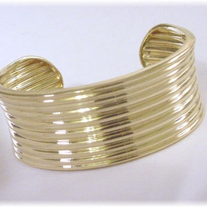 May include: A gold-toned bangle bracelet with a ridged design. The bracelet is made of multiple layers of metal that are stacked on top of each other.
