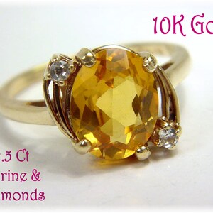Peut inclure: Une bague en or 10K avec une grande citrine jaune taille ovale au centre, entourée de deux petits diamants transparents. Le texte 10K Gold et 2.5 Ct Citrine & Diamonds est imprimé en rose sur l'image, soulignant les détails des matériaux.