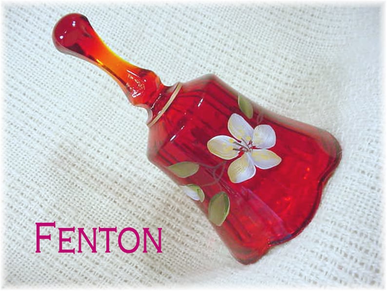 May include: A red glass bell with a white flower design. The bell is made by Fenton.