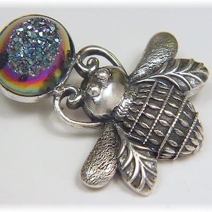 May include: A silver bee charm with iridescent gemstone accents. The bee has detailed wings and a textured body. The gemstone is round and has a blue, purple, and green iridescent sheen.