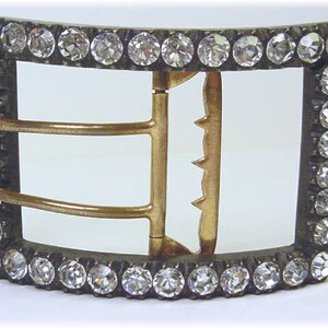 May include: A silver-toned buckle with a decorative design of clear rhinestones around the edge. The buckle has three gold-toned bars for attaching a strap.
