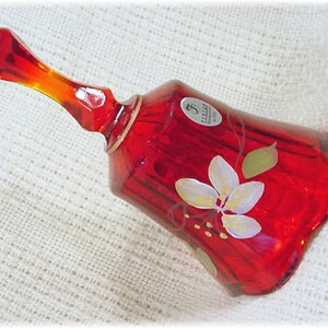 May include: A red glass bell with a white floral design and gold trim. The bell has a clear glass handle and a label that reads "F.  Schmid  Hand Painted".
