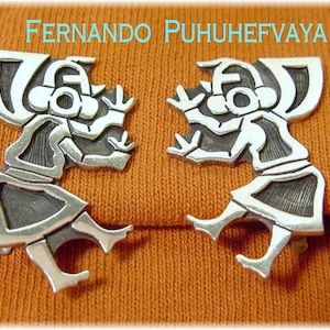 May include: Two silver-toned metal earrings with a stylized figure design. The earrings are in the shape of a person with a head, arms, and legs. The background is a textured orange fabric. The text "Fernando Puhuhefvaya" is at the top.
