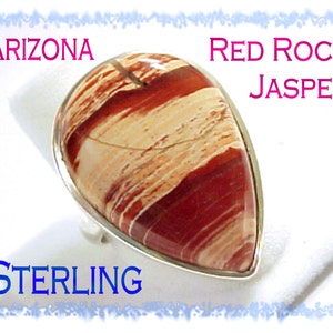 May include: A teardrop-shaped Arizona Red Rock Jasper gemstone set in a polished sterling silver ring. The stone features horizontal bands of deep red, terracotta, and cream colors. The text ARIZONA RED ROCK JASPER and STERLING is visible.