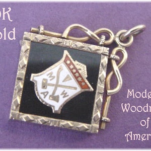May include: A gold-colored pendant with a black enamel square featuring a white emblem with the letters "MWA" and a hammer and axe. The pendant is attached to a gold-colored chain with a decorative loop. The text "10K Gold" and "Modern Woodman of America" are visible in the image.