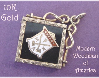 May include: A gold-colored pendant with a black enamel square featuring a white emblem with the letters "MWA" and a hammer and axe. The pendant is attached to a gold-colored chain with a decorative loop. The text "10K Gold" and "Modern Woodman of America" are visible in the image.
