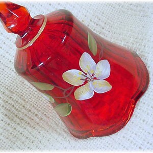May include: A red glass bell with a white flower painted on it. The bell has a scalloped edge and a gold rim.
