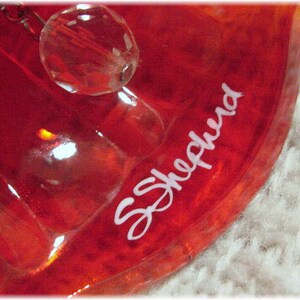 May include: A red glass ornament with a clear crystal hanging from it. The ornament has the name "S.Shepnerd" written on it in white.
