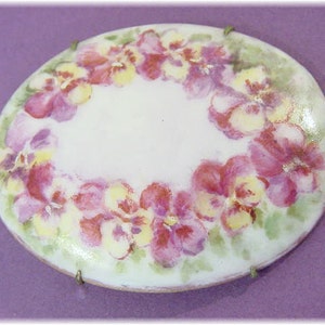 Puede incluir: Broche ovalado con un diseño floral pintado a mano. El broche presenta un centro blanco rodeado por una corona de pensamientos rosas y amarillos sobre un fondo verde pálido. El broche se asegura con pequeños cierres metálicos.